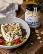 Ser feta z liofilizowanym kiszonym czosnkiem Johny Catch, oliwą, oliwkami i pomidorkami koktajlowymi, obok słoiczek produktu.