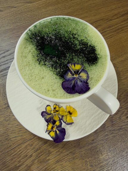 Filiżanka napoju Matcha + Pacific Johny Catch z zieloną pianką, proszkiem spiruliny pacyficznej i jadalnymi liofilizowanymi bratkami na białym spodku – naturalny, roślinny napój superfood.