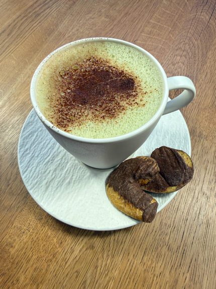 Filiżanka napoju Matcha + Cacao Johny Catch z aksamitną pianką i posypką z kakao, podana na białym spodku z kawałkami liofilizowanej śliwki z czekoladą – naturalna energia i słodycz w jednym.