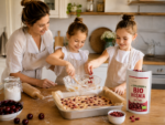 Mama z dwiema córkami pieką domowe ciasto z wiśniami w jasnej kuchni. Dziewczynki dodają do masy liofilizowane BIO wiśnie Johny Catch, a obok na blacie stoi otwarte opakowanie produktu, miseczka z wiśniami i forma z przygotowanym plackiem.